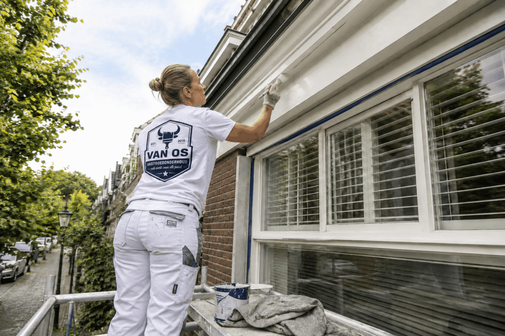Schilder van Van Os op de steiger schildert een witte dakrand boven een erker tijdens buitenschilderwerk in Amsterdam