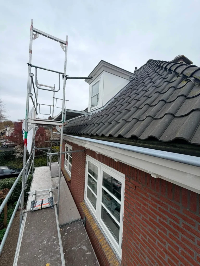 Steiger langs pannendak en bakstenen woning met witte kozijnen tijdens dakrenovatie